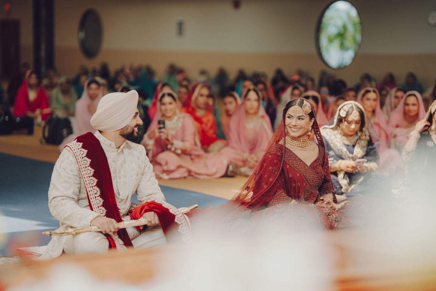 Traditional Sikh wedding ceremony showing guests seated in rows at a Gurdwara with red and pink attire.