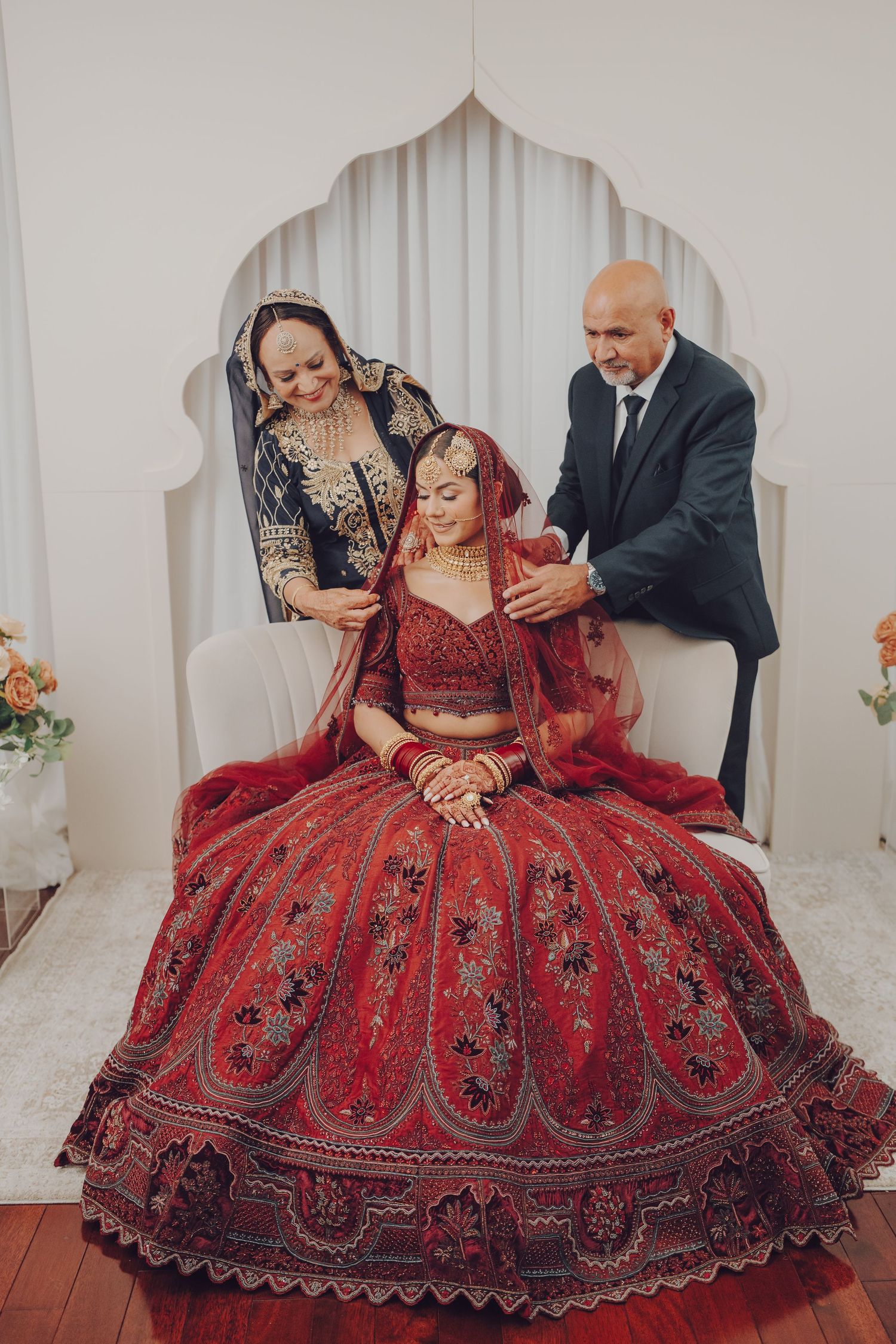 Traditional Indian wedding ceremony with family members gathered around bride in ornate red lehenga under arched backdrop.