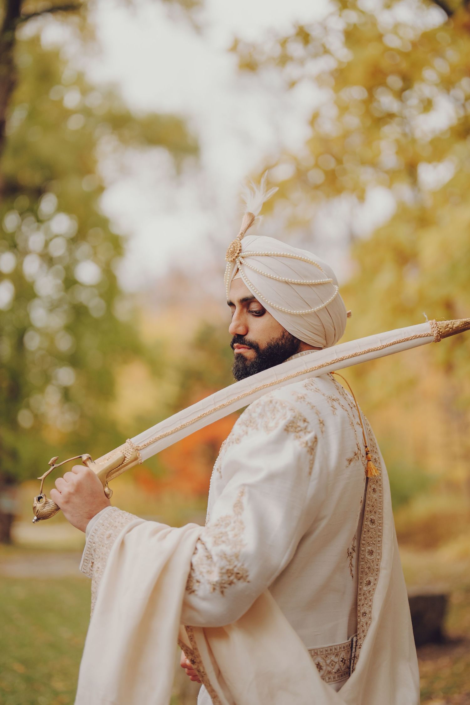 Artistic side view of traditional Sikh groom in cream outfit against autumn foliage.