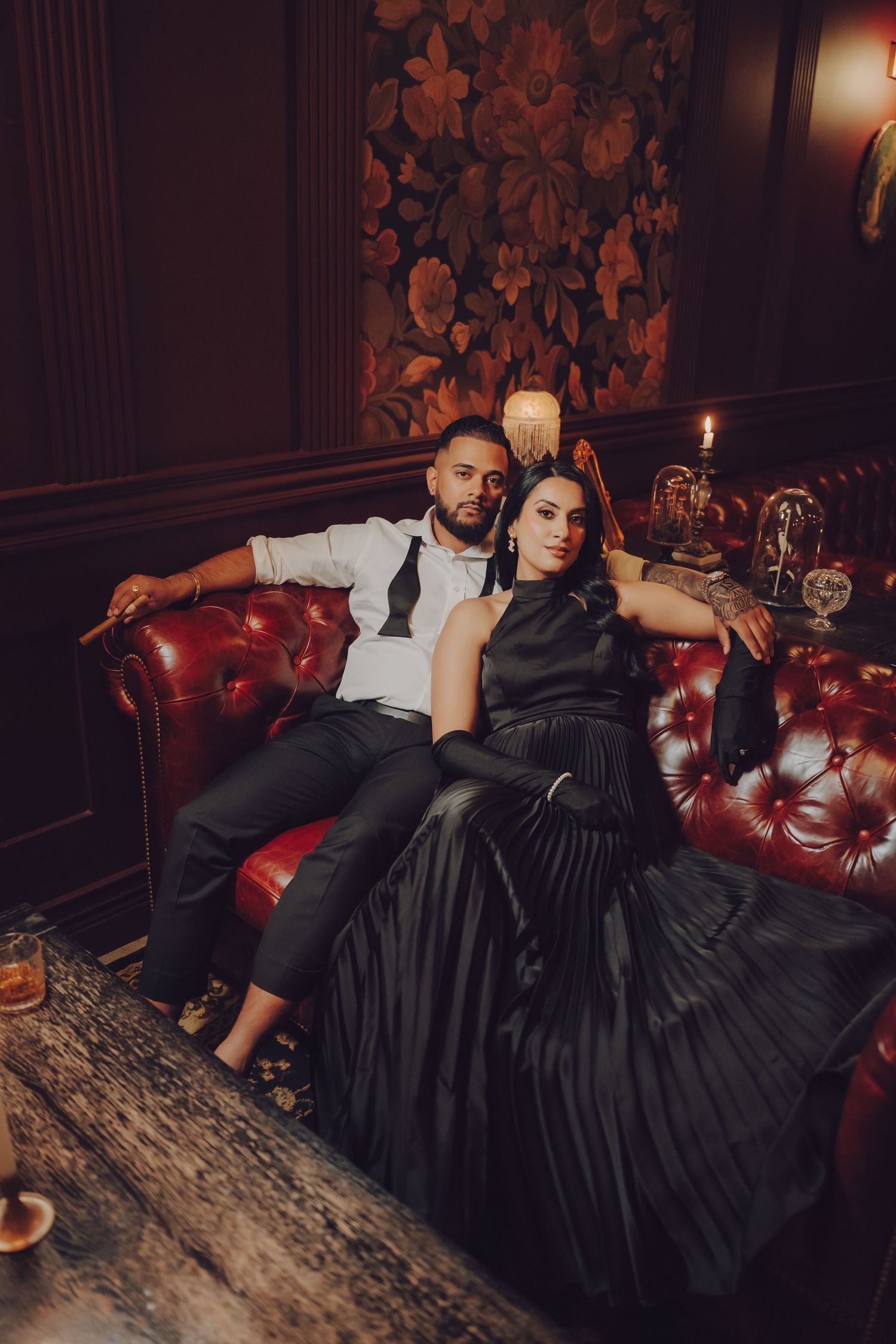 Elegant couple lounging on a vintage red leather chesterfield sofa in formal evening attire.