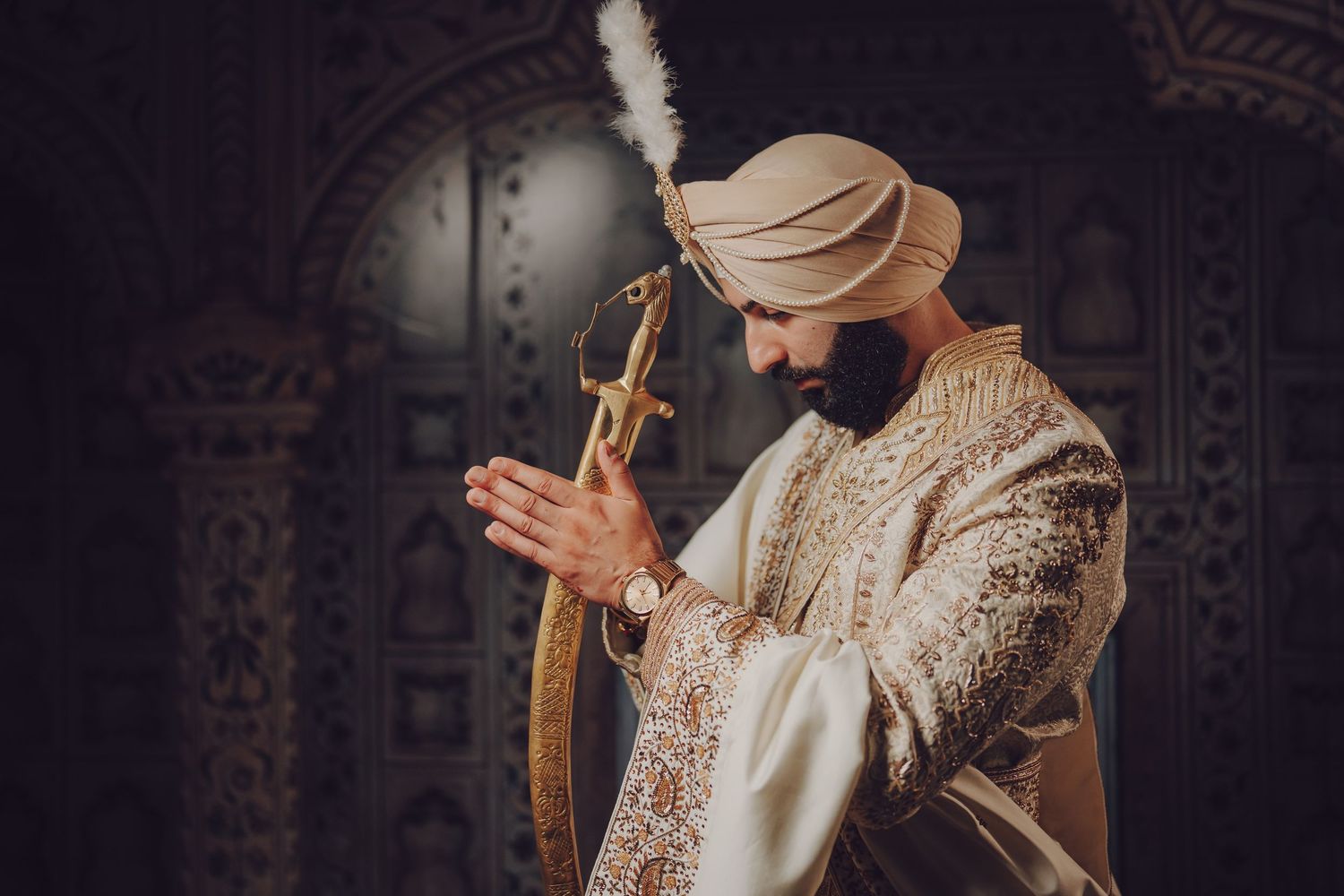 Groom in ornate Sikh wedding attire holding ceremonial sword against patterned backdrop.