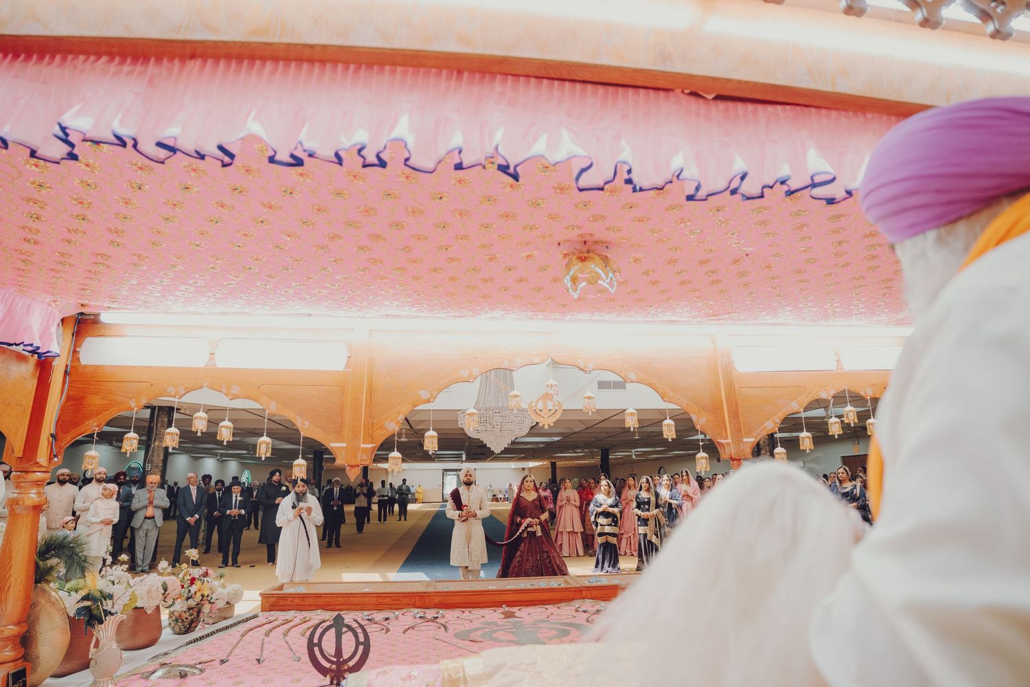 Interior view of Sikh wedding ceremony with guests gathered under ornate pink ceiling.