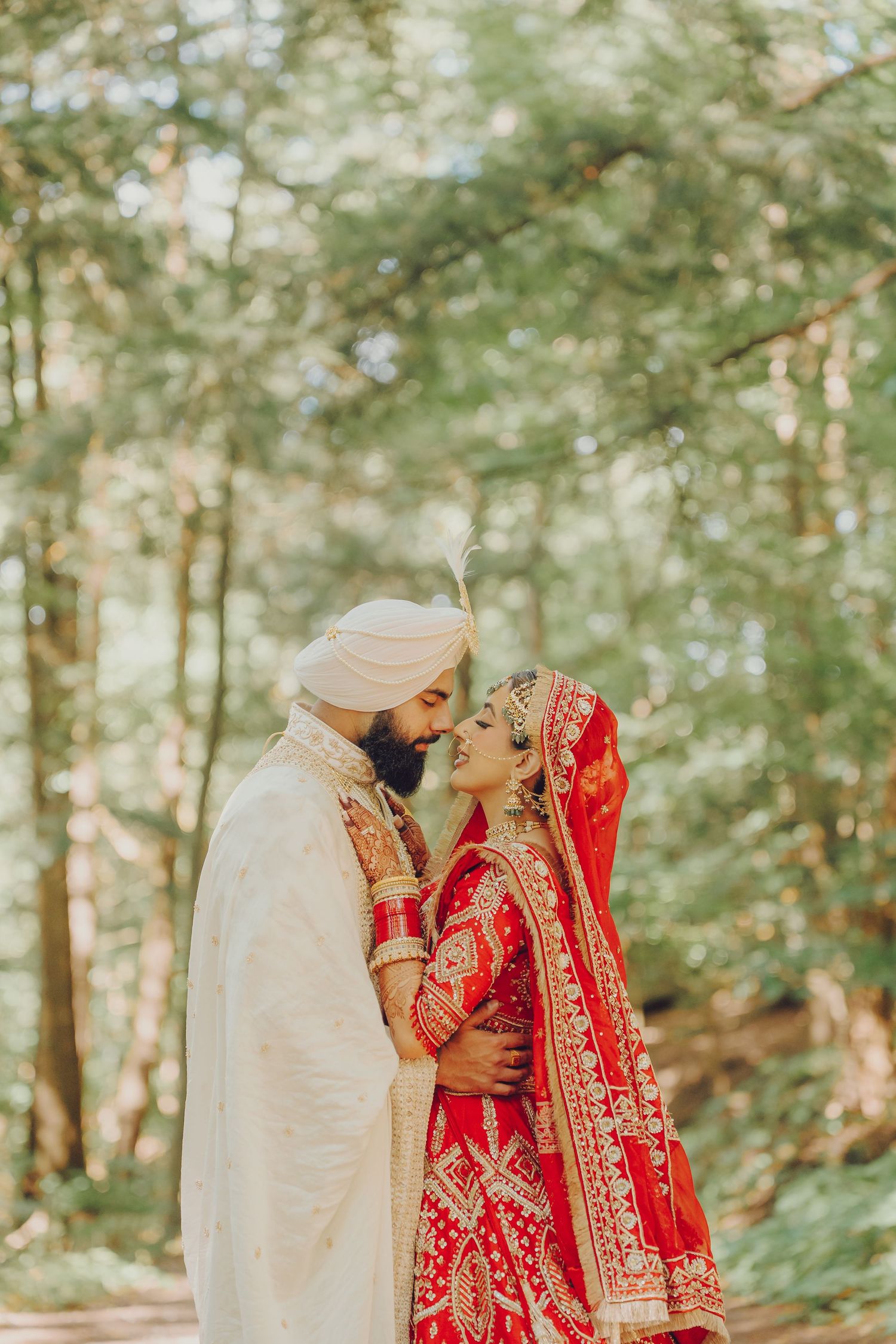 Traditional Indian wedding couple in red and white attire posing together in a wooded outdoor setting.