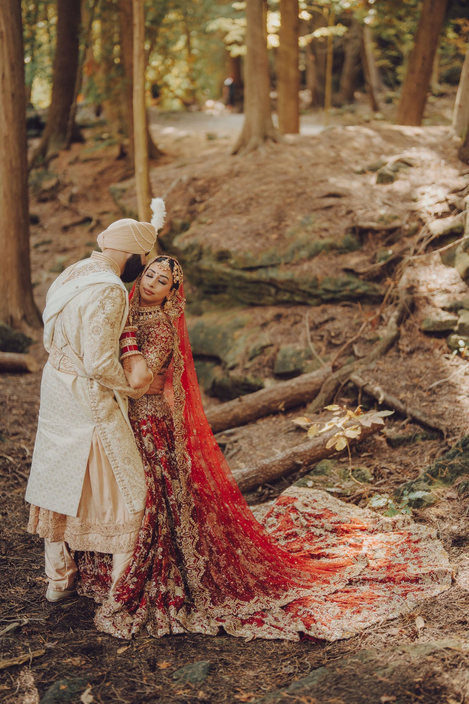 A couple in traditional wedding attire poses in a wooded forest setting with a flowing red bridal dress train.