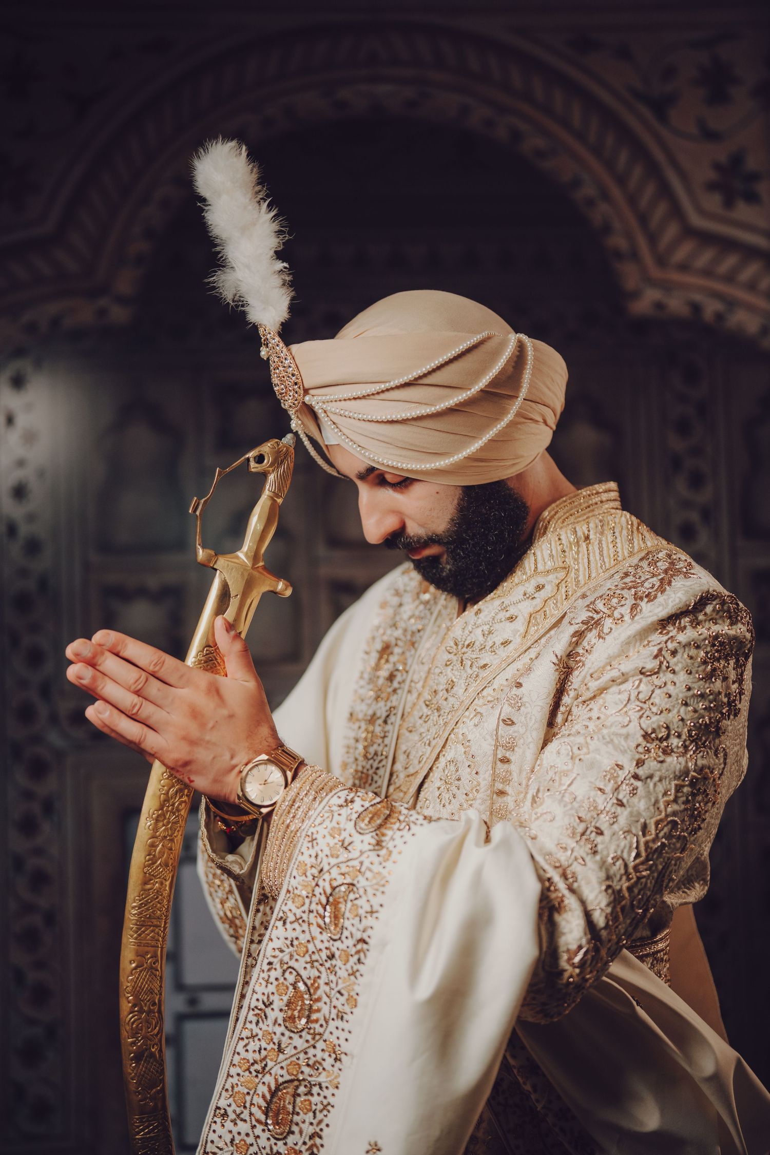 A groom in cream and gold attire with turban and feather accent prays in traditional style.