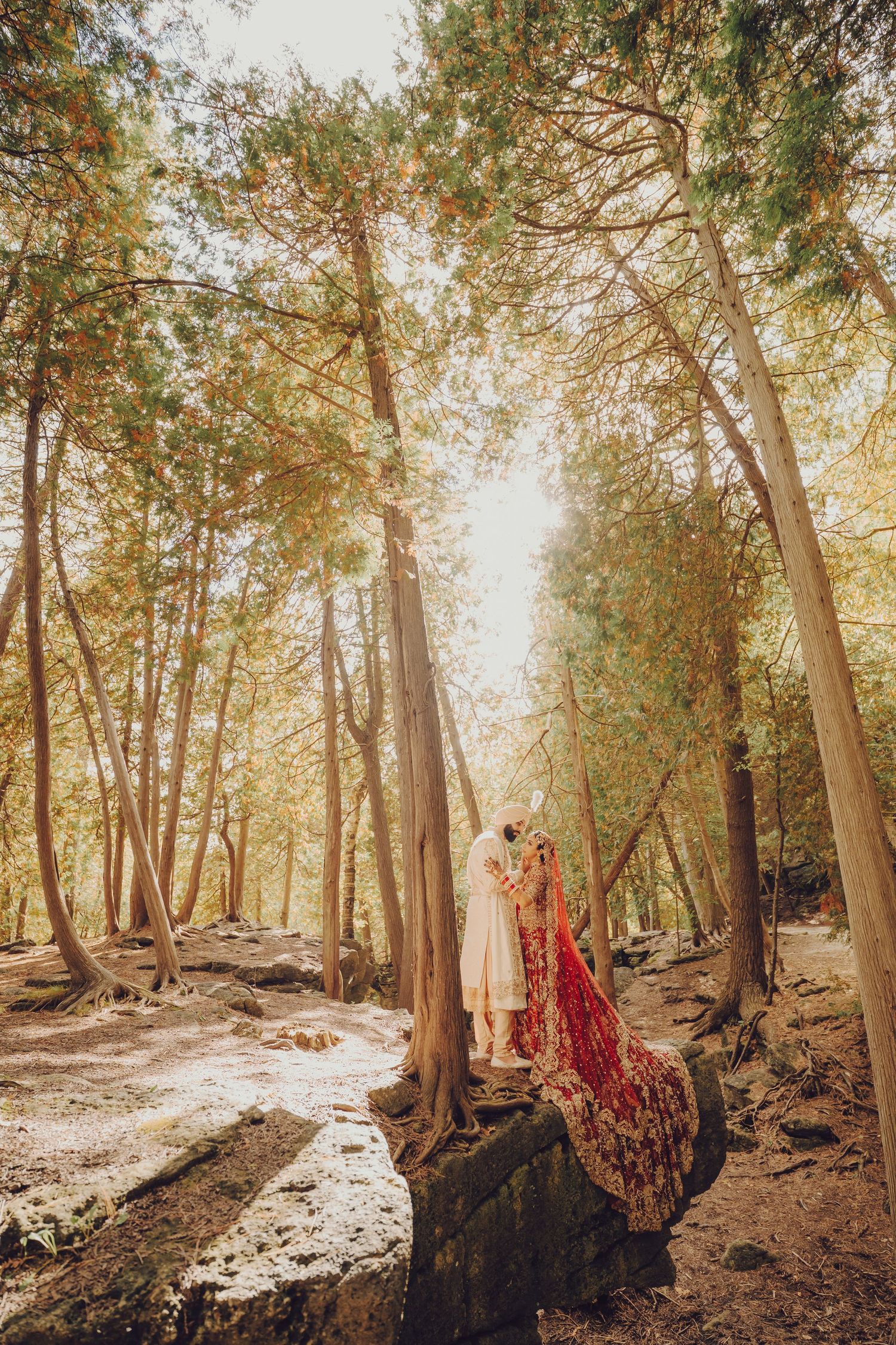 A couple in traditional Indian wedding attire stand together in a sunlit forest setting with a flowing red dupatta.