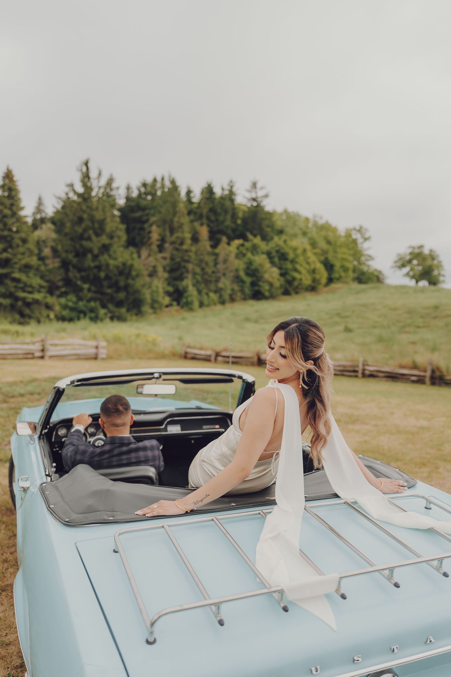 Playful couple poses in vintage light blue convertible car on a hilltop.