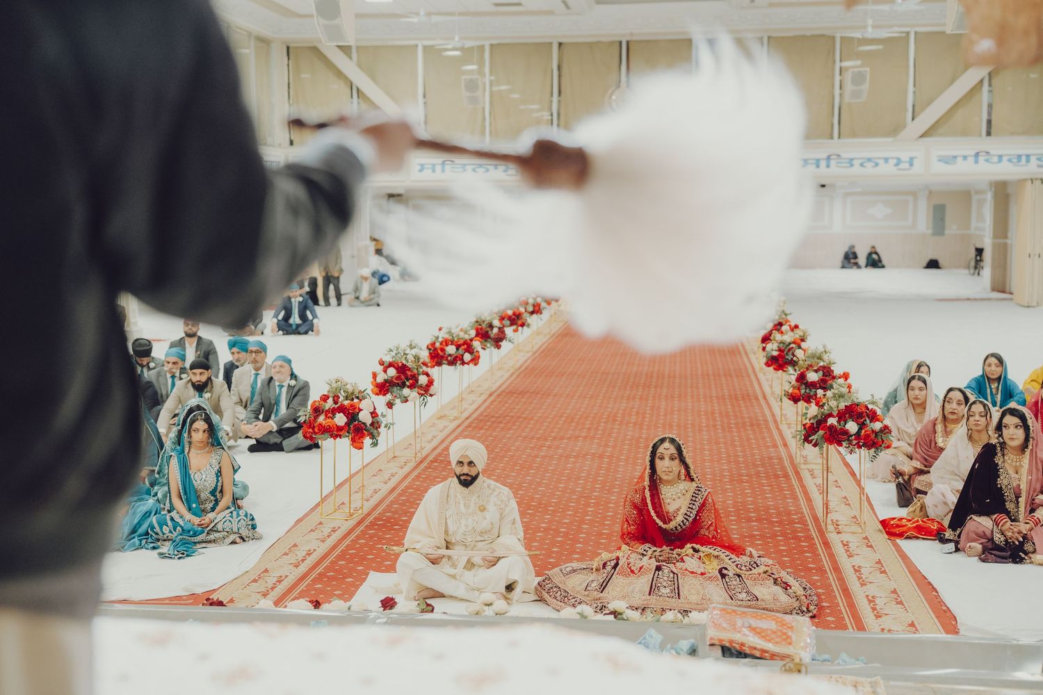 Wedding guests seated on floor during traditional Sikh ceremony in decorated indoor venue.