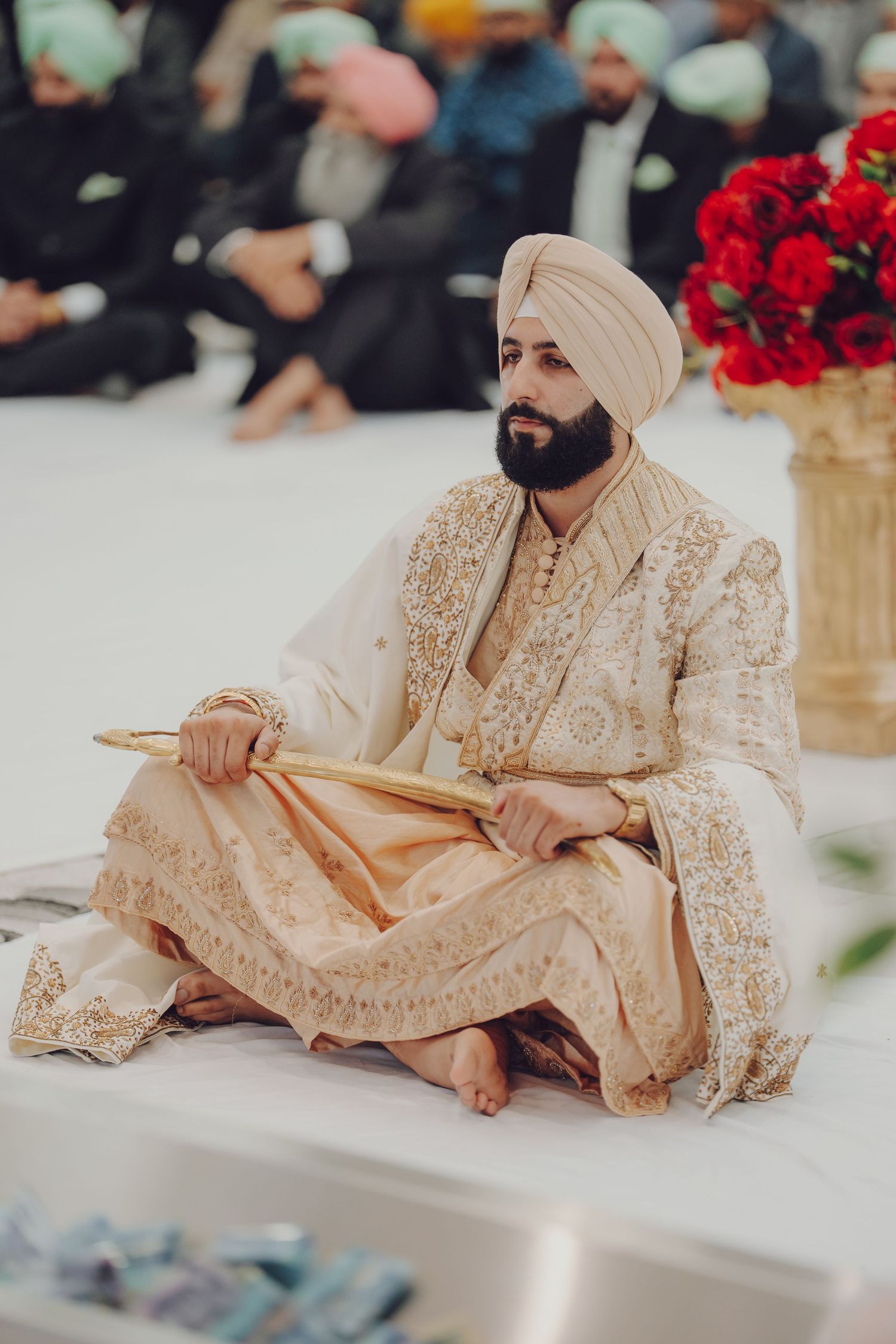 Traditional Sikh wedding ceremony with attendees in ornate cream-colored outfits seated on white floor.