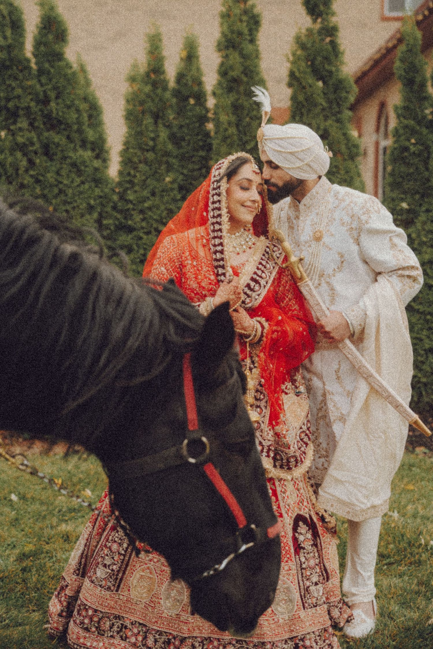 A couple in traditional Indian wedding attire poses with a black horse during an outdoor photo session.