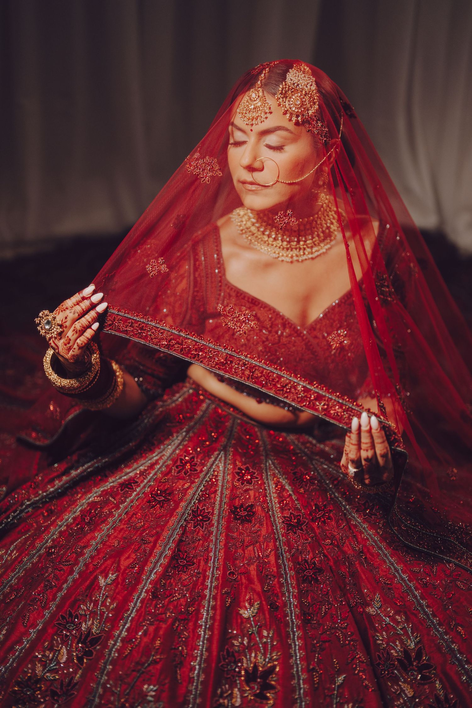 Traditional Indian bride in ornate red lehenga with detailed embroidery and veil poses elegantly.