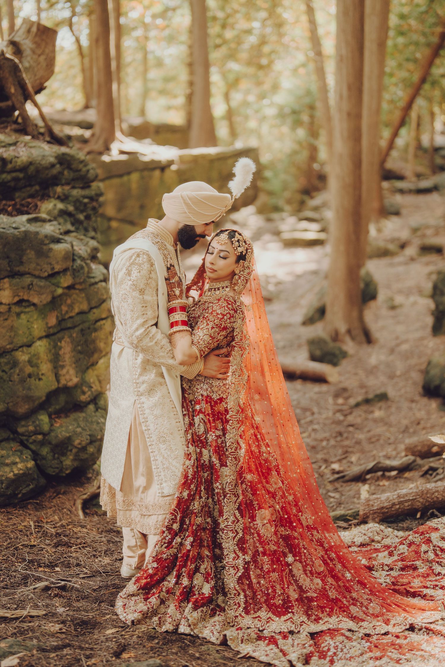 Traditional South Asian wedding couple poses in a forest setting wearing ornate red and cream ceremonial attire.