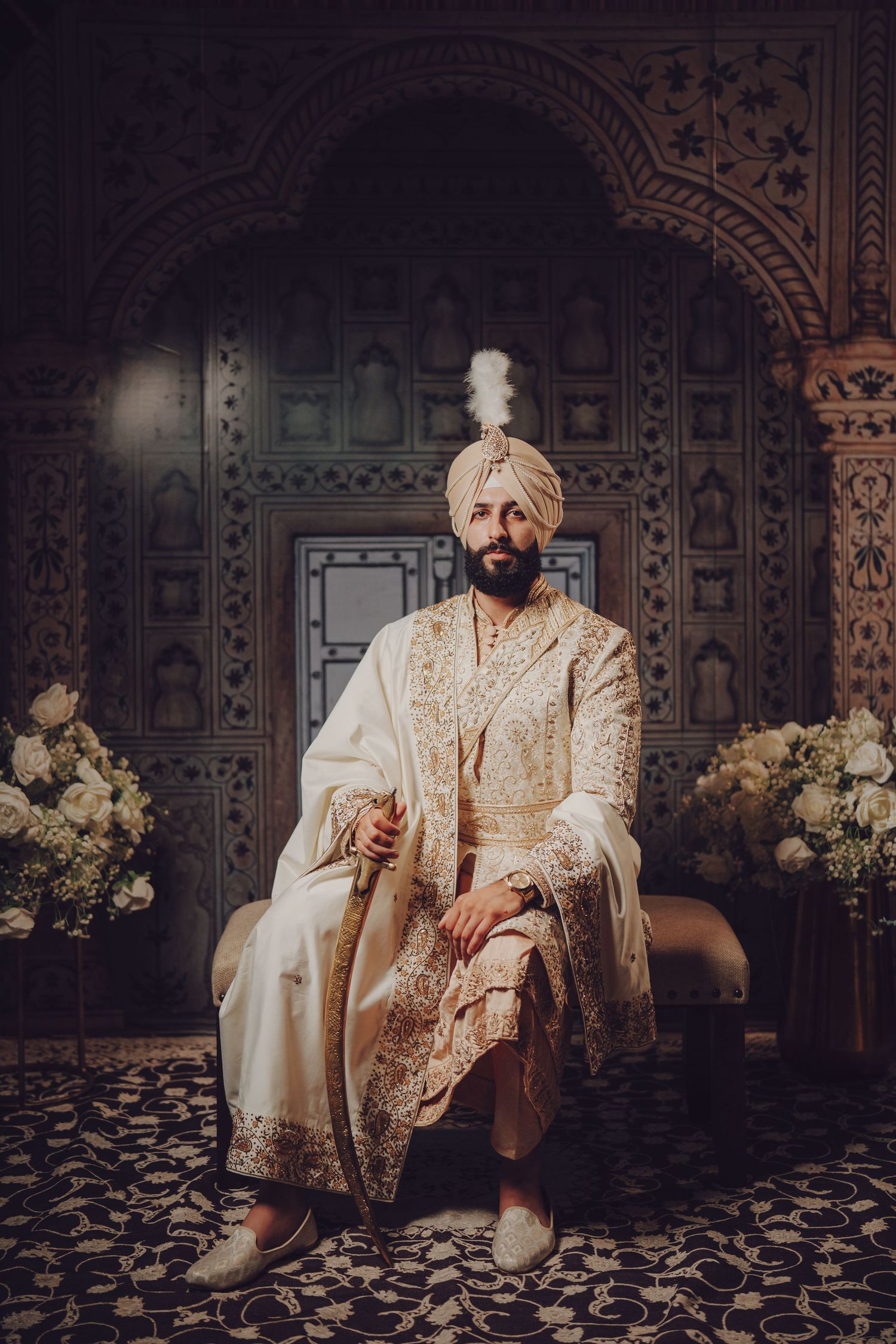 Traditional Indian groom in cream and gold sherwani and turban seated in front of ornate carved backdrop.
