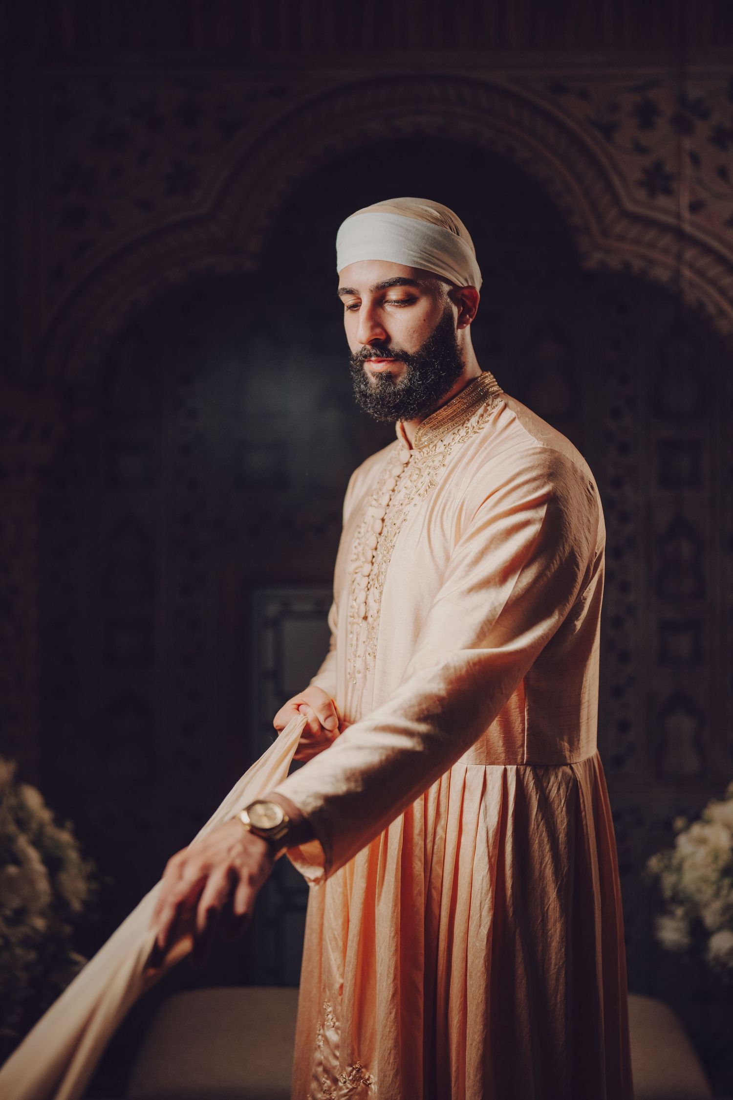 Groom in traditional cream-colored Indian wedding attire poses against ornate architectural backdrop.