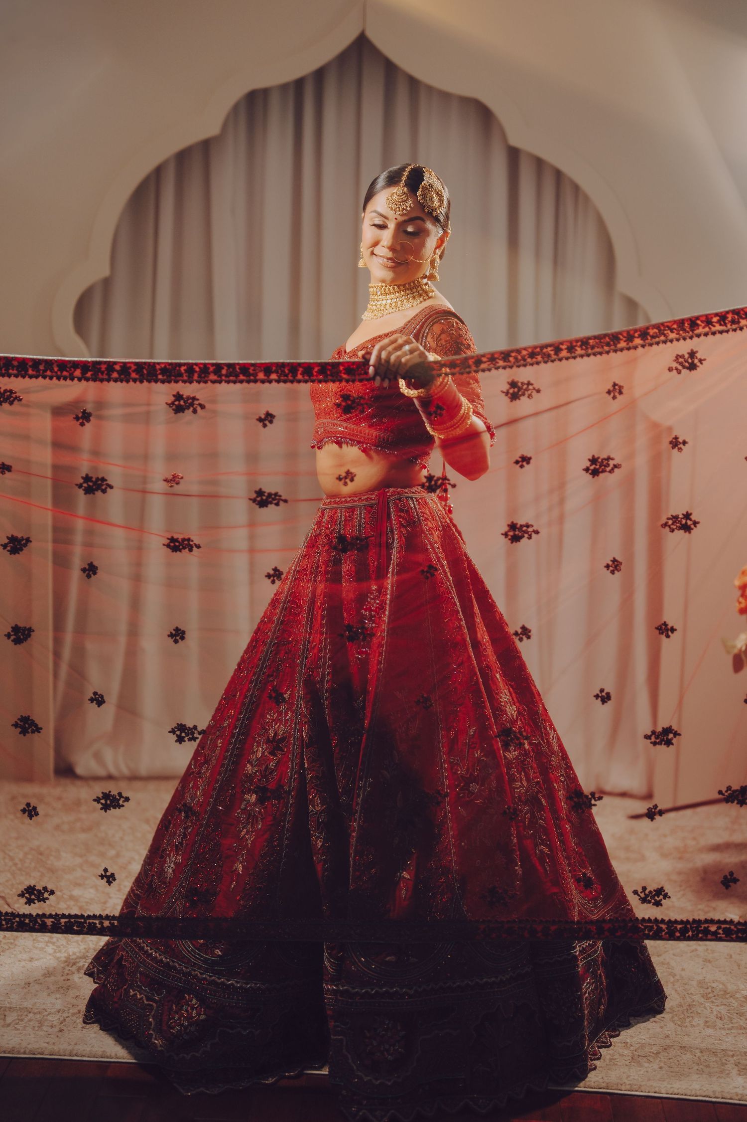 Bride in stunning red embroidered lehenga poses gracefully against white backdrop with arched design.