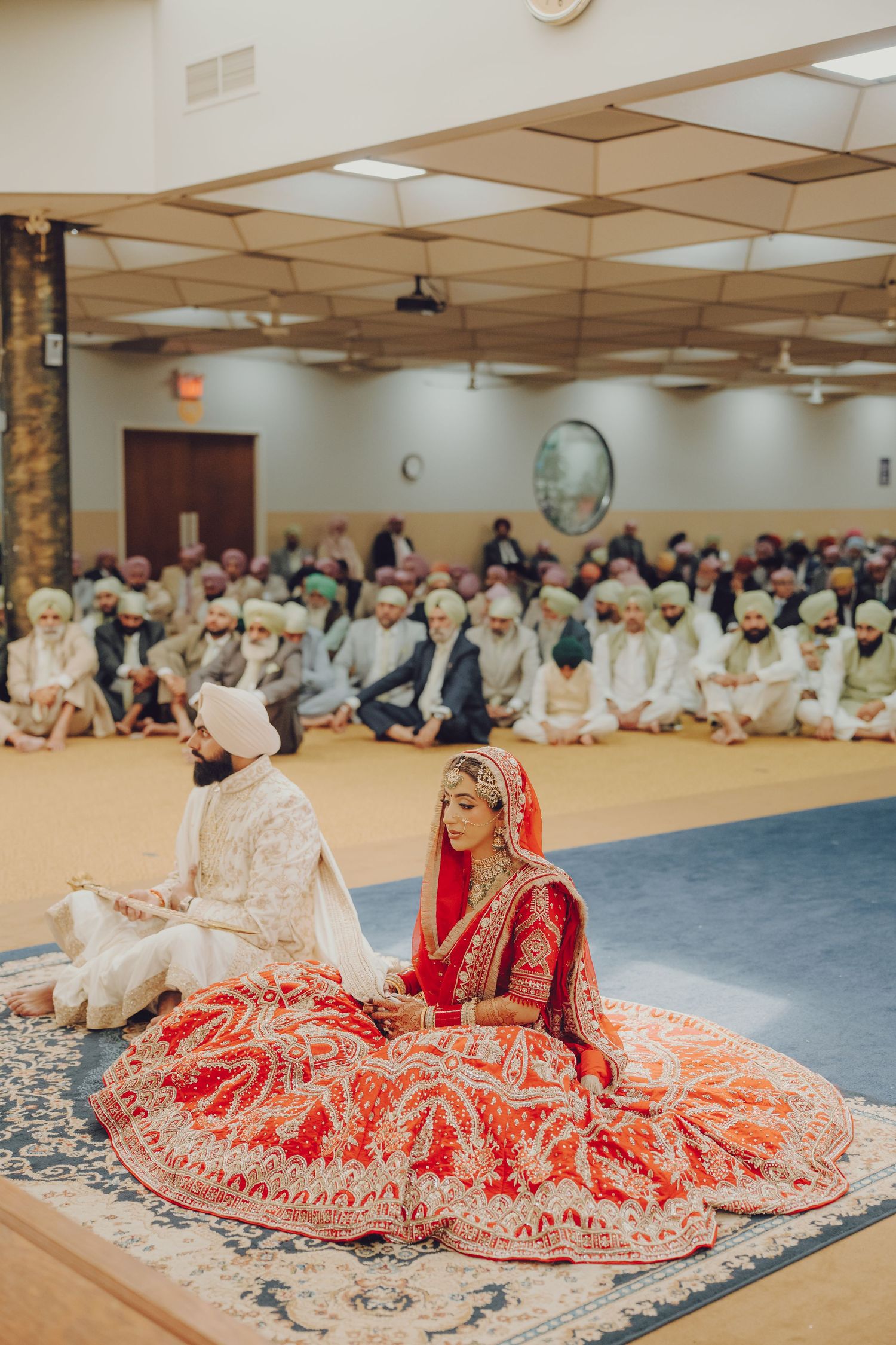 Wedding ceremony in community hall with couple in traditional attire seated before gathering.