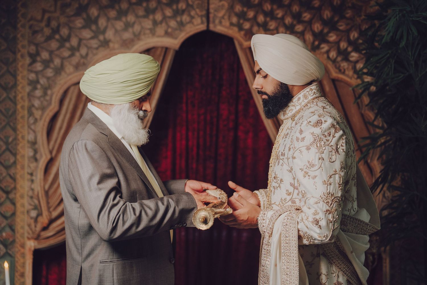 A couple in traditional Indian wedding attire holds hands during a ceremony in an ornately decorated venue.