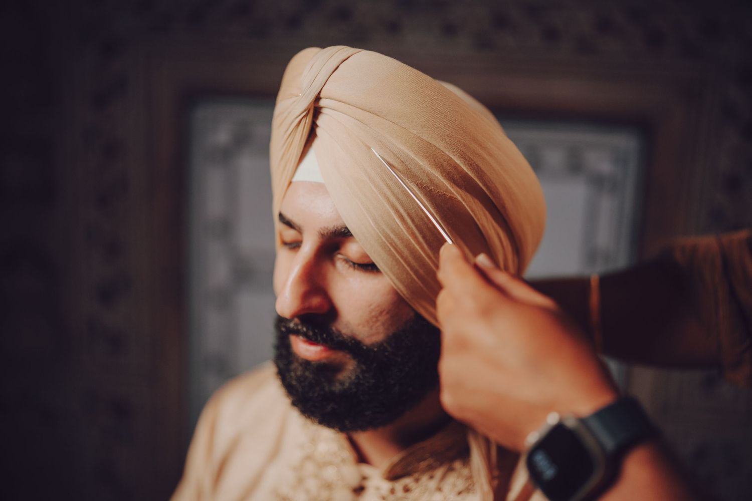 A traditional Sikh turban being tied in a dimly lit room with ornate decor.