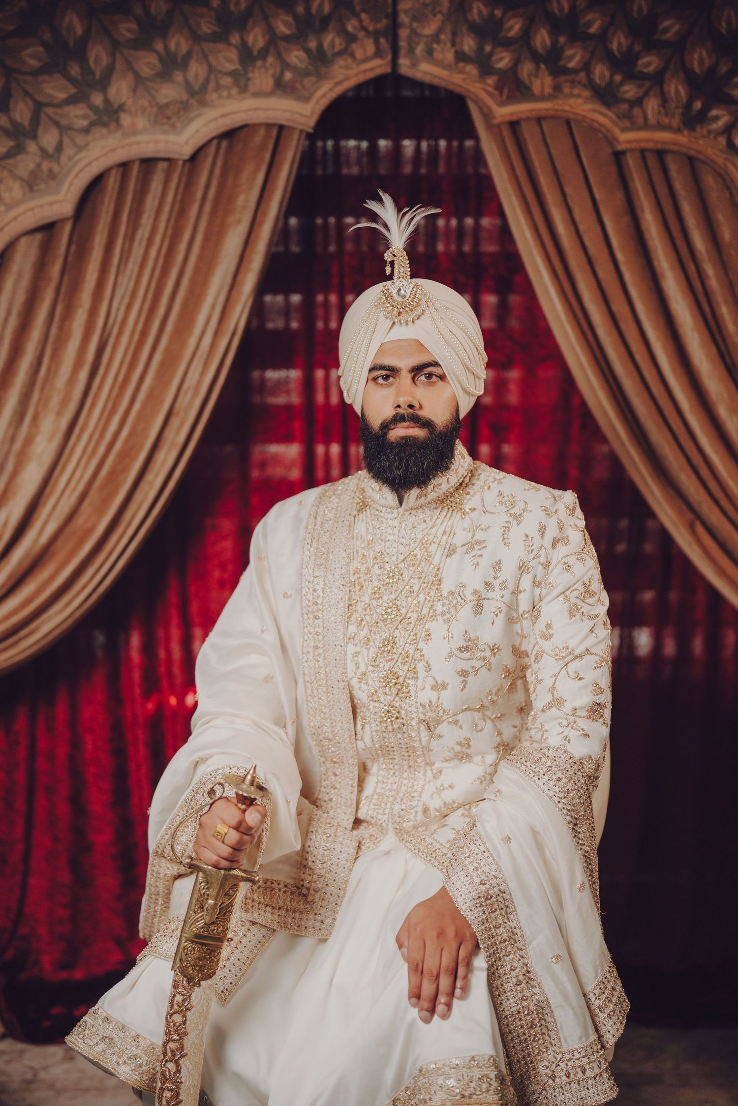 Traditional Sikh groom in white sherwani and turban holding ceremonial sword against red curtains.