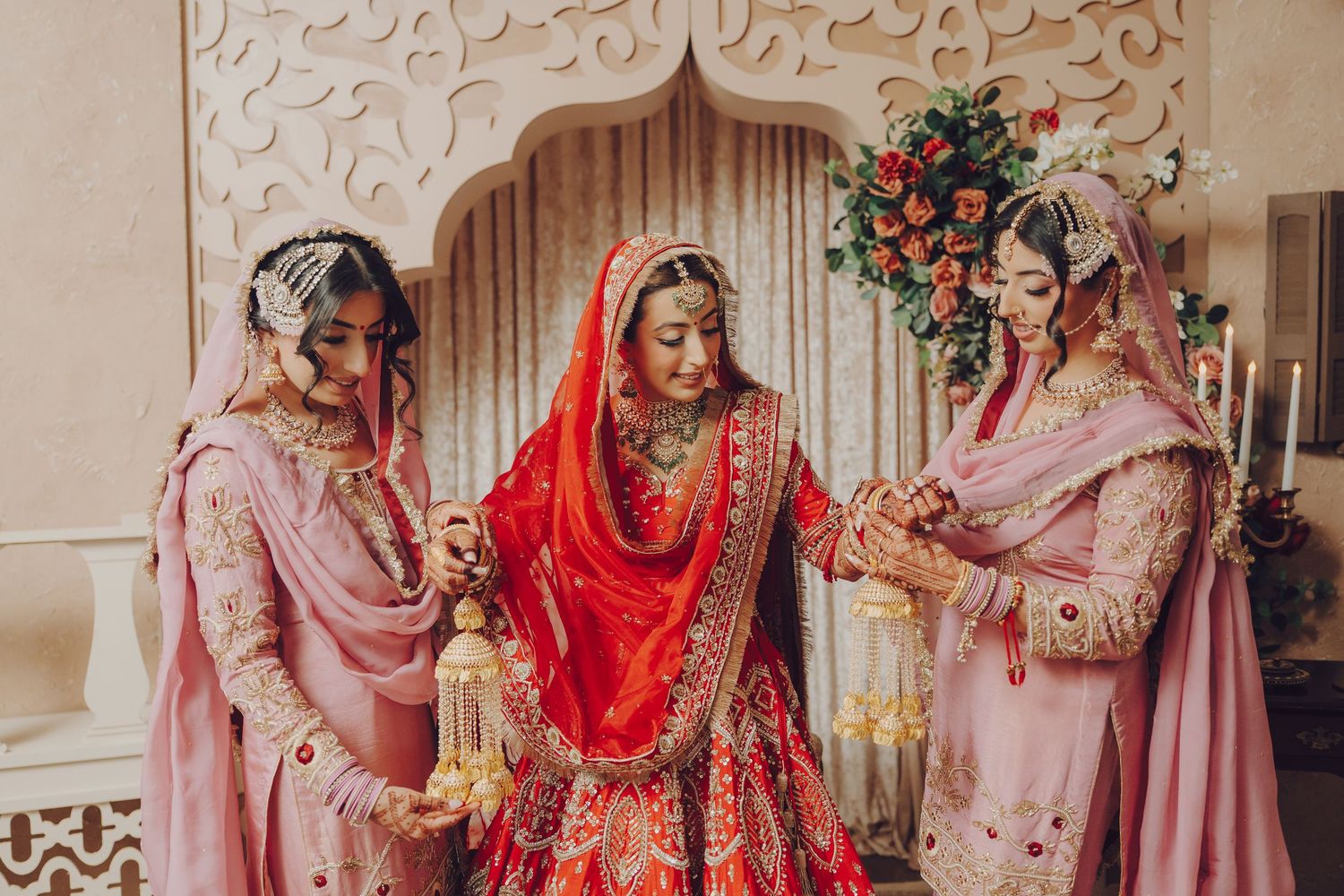 Three women in traditional pink and red Indian wedding attire standing together during a ceremony.