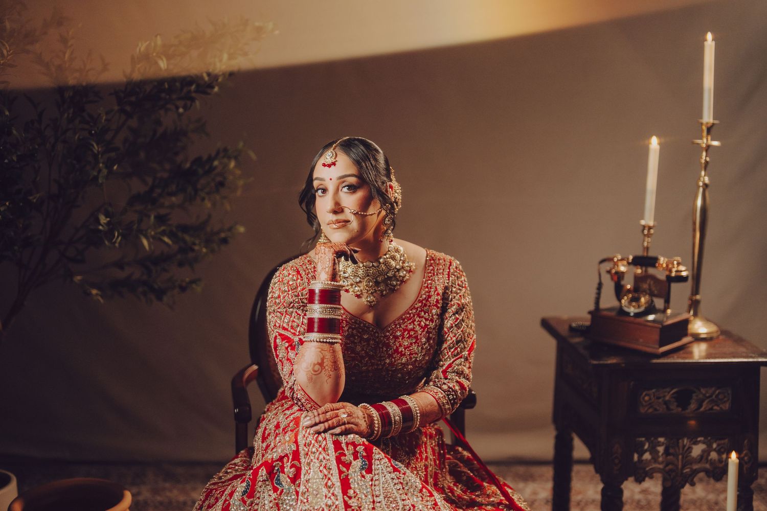 Traditional Indian bride in ornate red lehenga poses elegantly beside antique table with candlesticks in moody lighting.