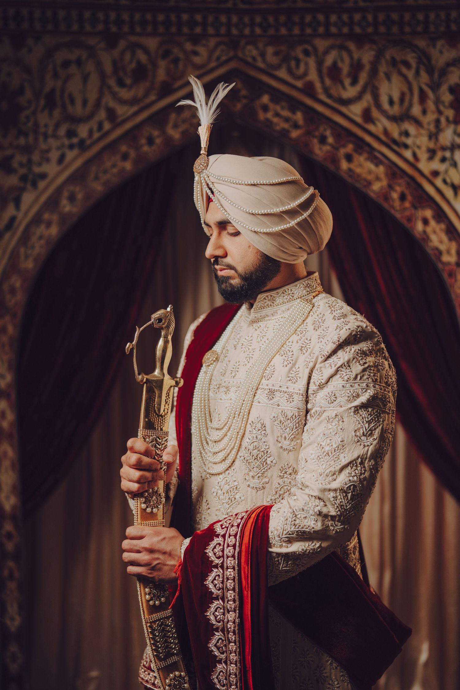 Sikh groom in cream sherwani and red dupatta holding ceremonial items during wedding ritual.