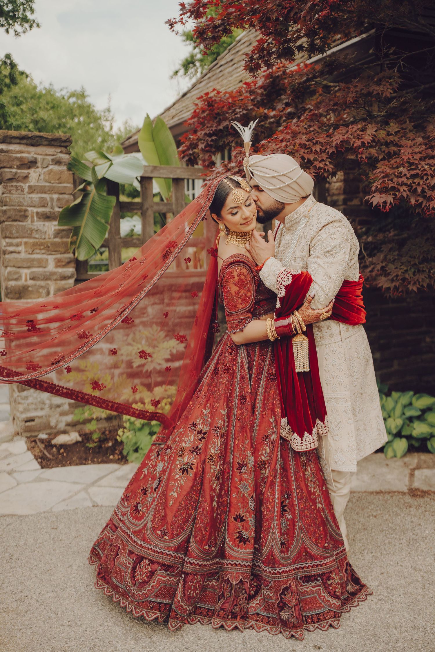 Wedding couple in red and white South Asian attire pose romantically against a backdrop of red foliage.