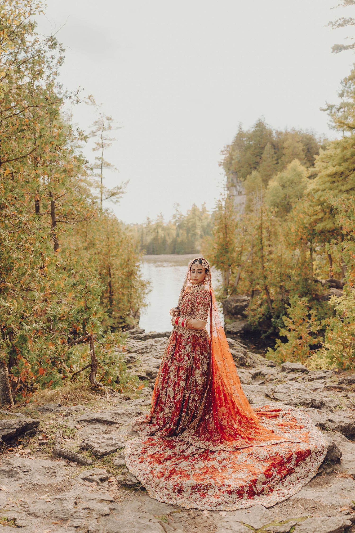 A bride in an ornate red lehenga with a flowing train poses on rocky terrain near a lake.