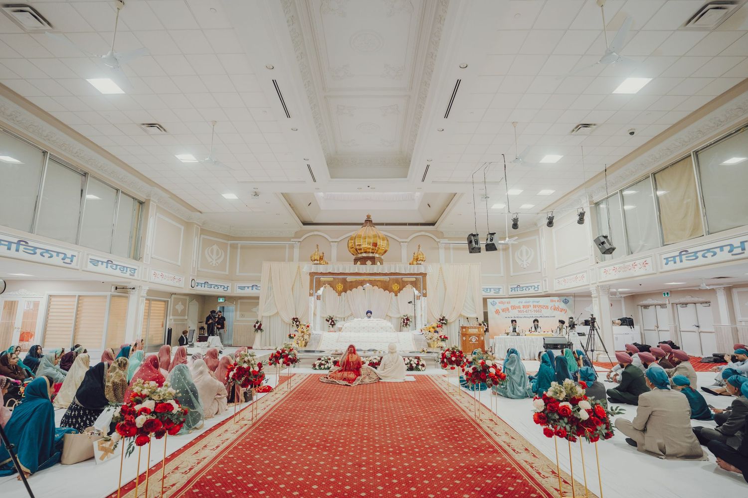 Wide panoramic view of a Sikh temple interior with red carpeting and seated guests.