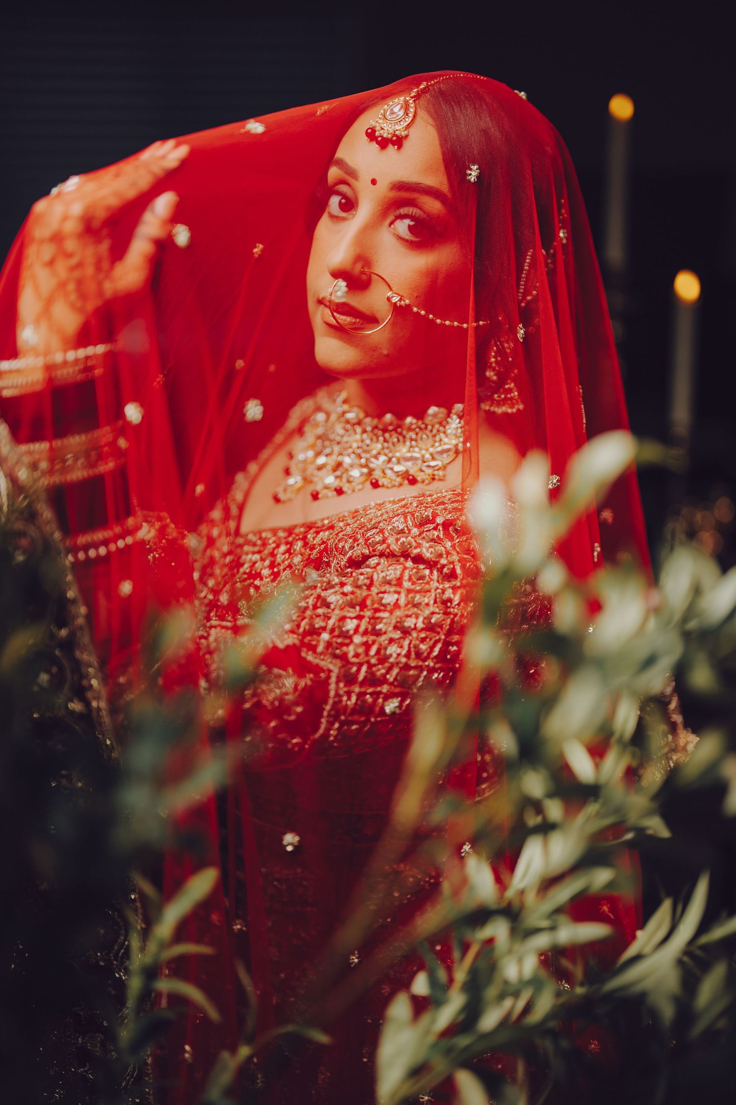 Traditional Indian bride in red attire with ornate jewelry and veil poses against blurred floral backdrop.