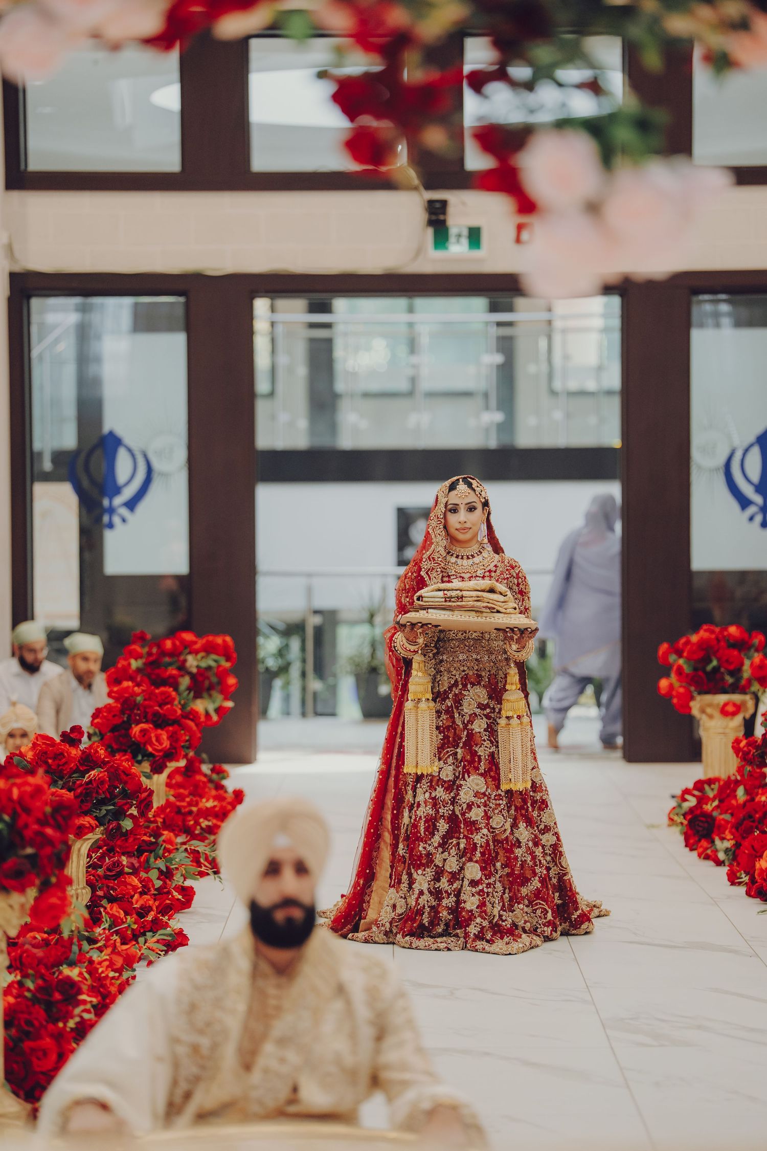 Traditional Sikh wedding ceremony with red floral decorations and ornate bridal attire in a modern venue setting.