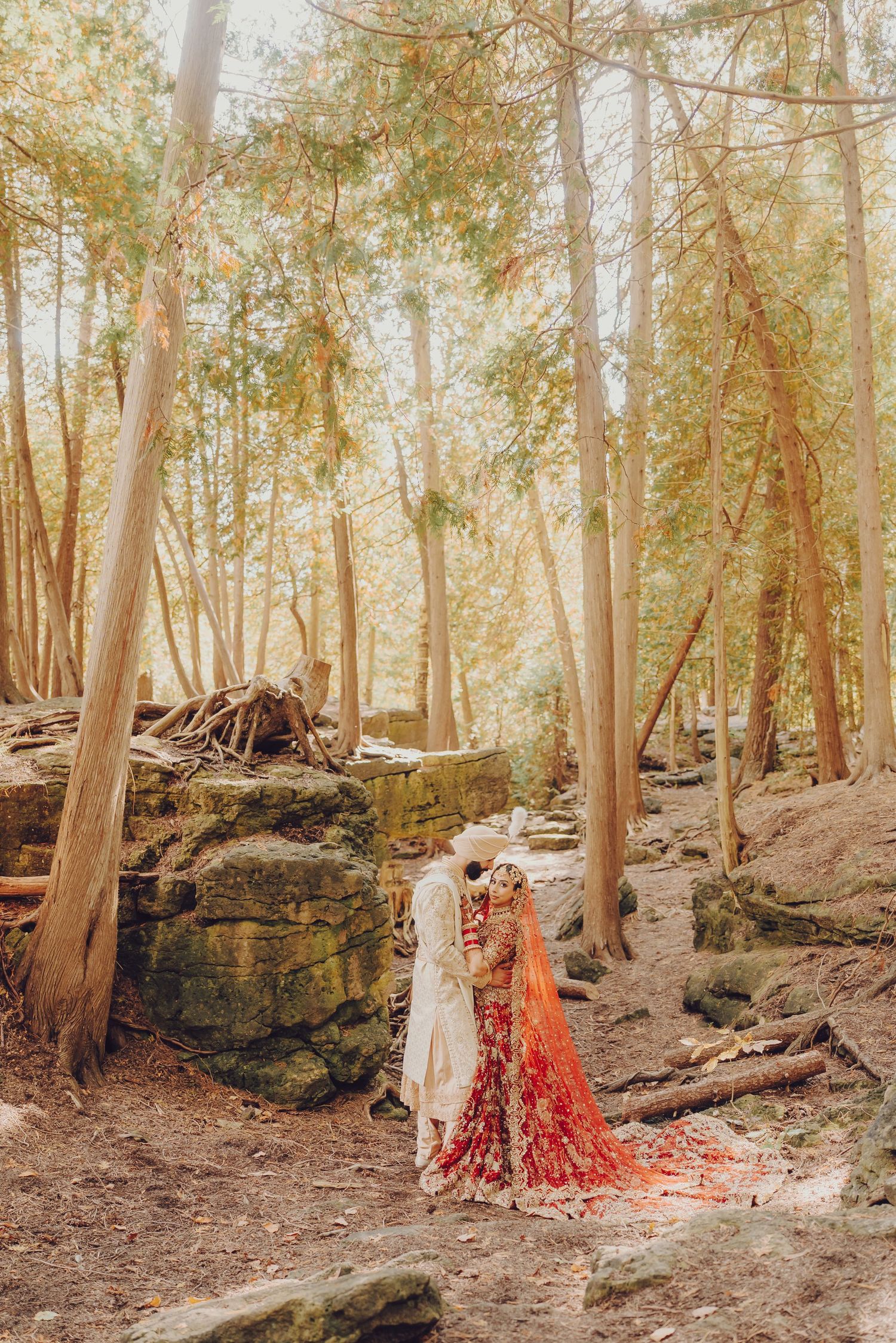 A couple in traditional wedding attire poses in a dreamy forest setting with sunlit trees and rocky terrain.