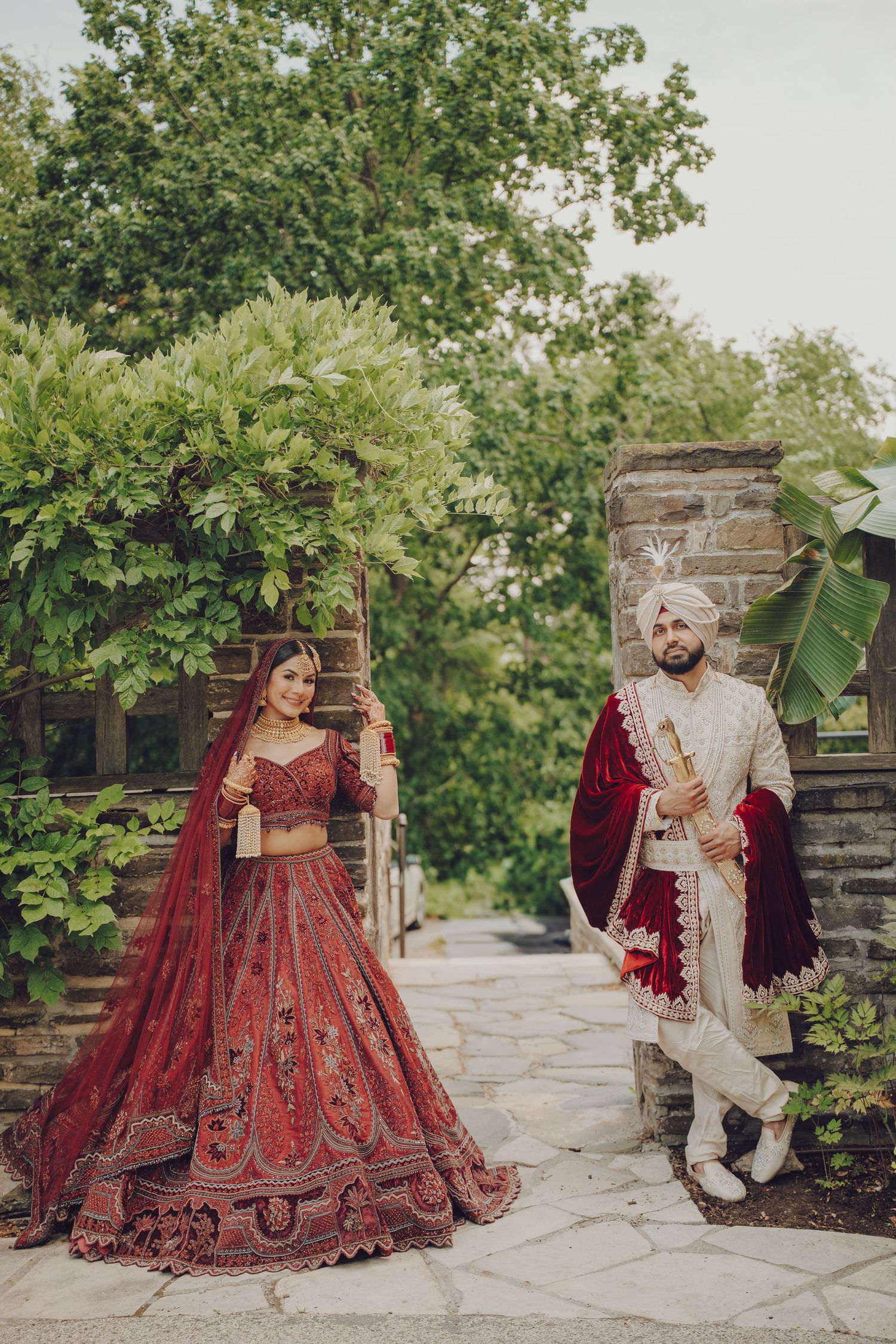 A couple in traditional South Asian wedding attire poses together in a garden setting with brick walls and greenery.