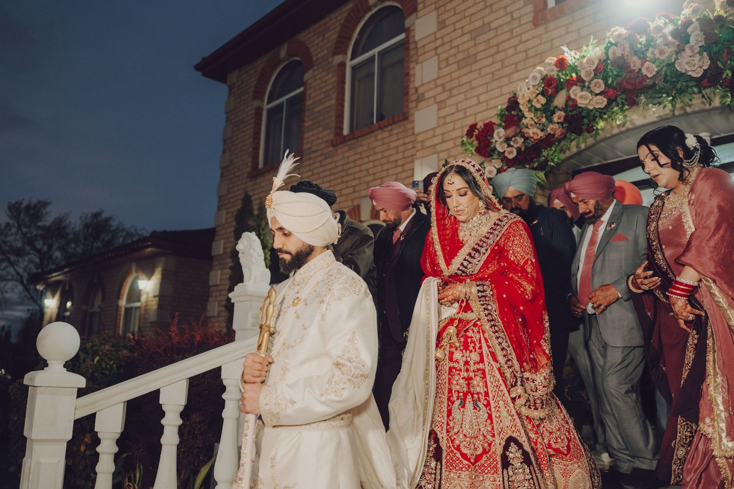 Traditional Indian wedding ceremony taking place outside a brick building at dusk with decorative lighting.