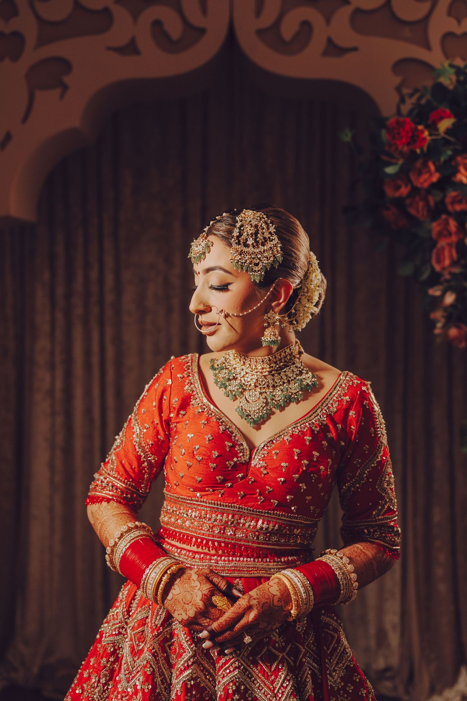 A traditional Indian bride in an ornate red lehenga poses elegantly against a dark backdrop with floral decorations.