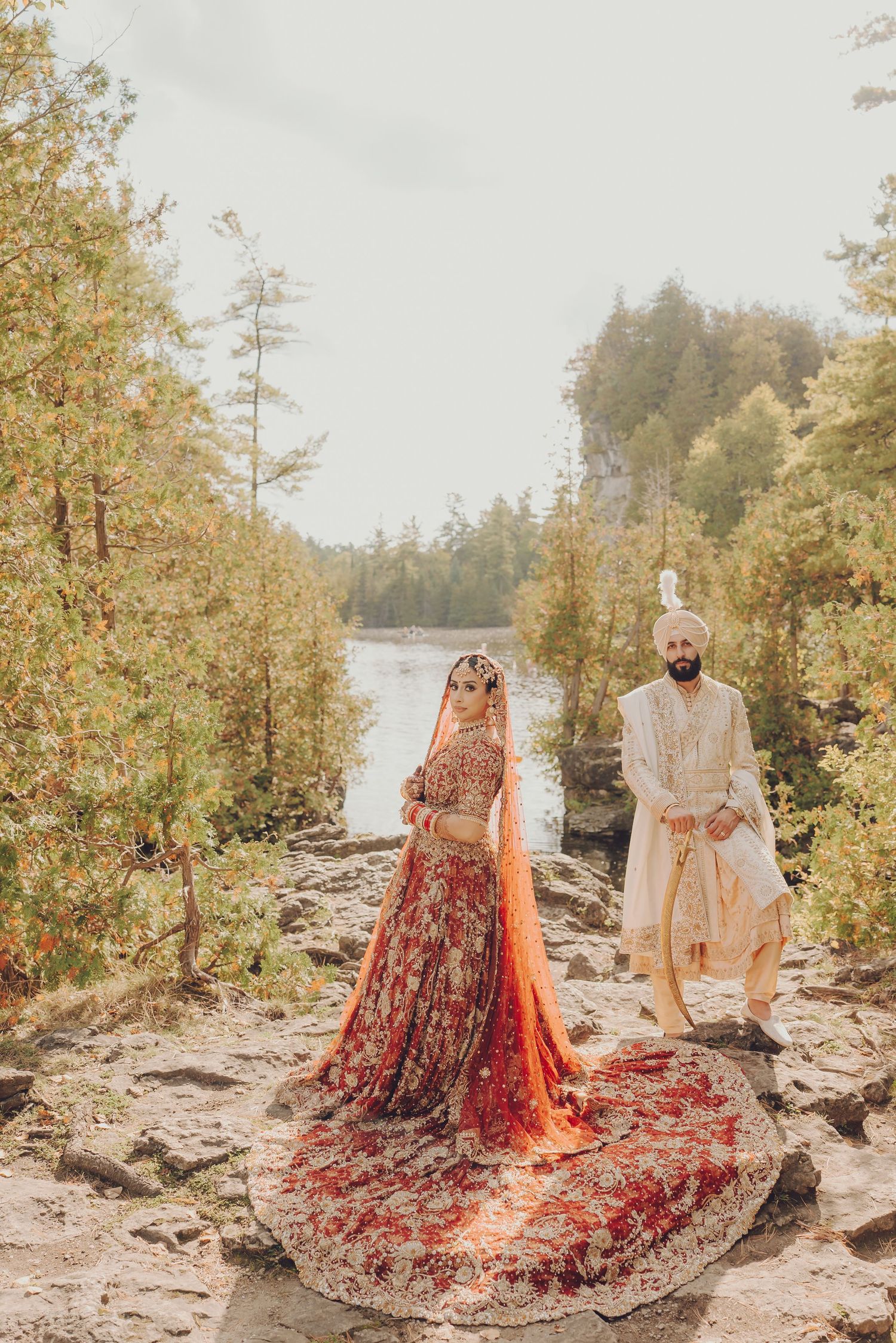 A couple in traditional South Asian wedding attire poses against a scenic lakeside backdrop with autumn foliage.