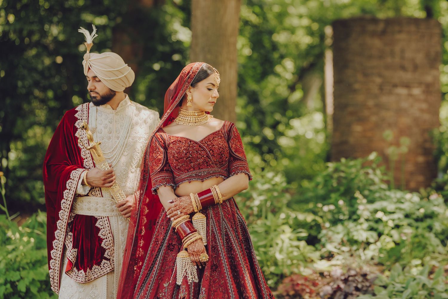 Couple in traditional Indian wedding attire walking through garden setting.