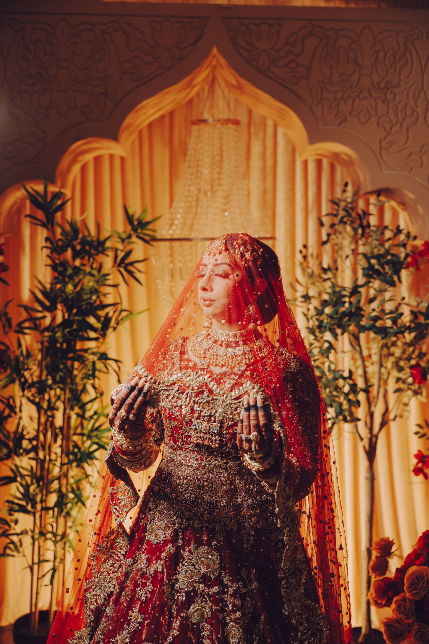 Traditional Indian bride wearing an ornate deep red lehenga and veil stands against illuminated archway backdrop.
