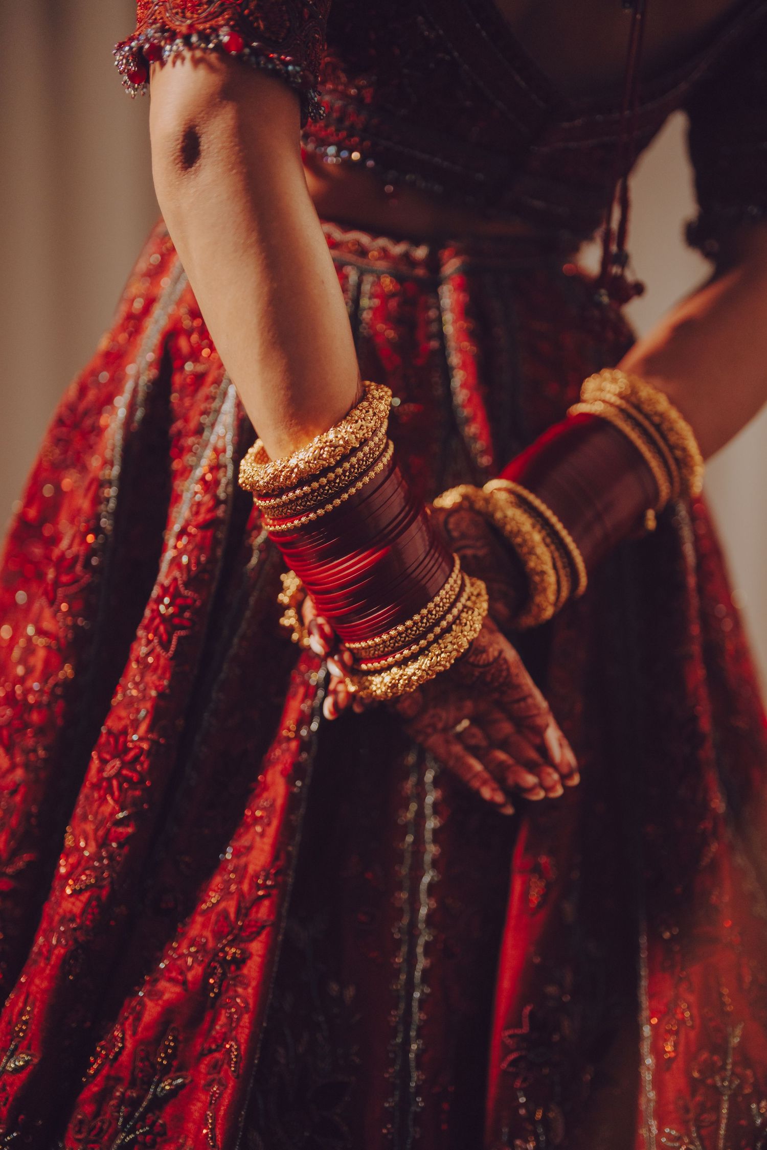 Close-up of intricate gold bangles and jewelry adorning hands against red bridal lehenga.