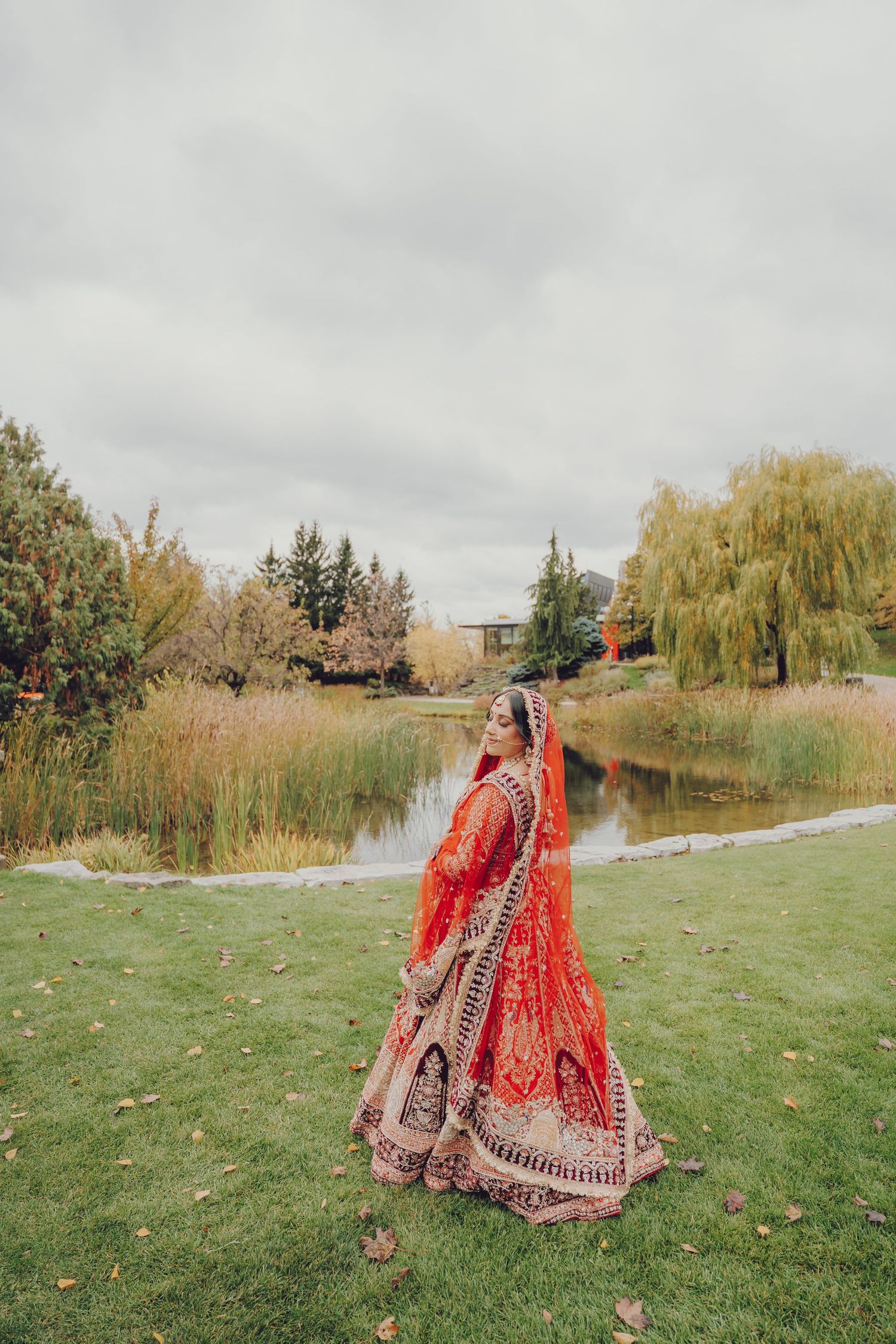 Bride in red lehenga standing in garden setting with autumn foliage.