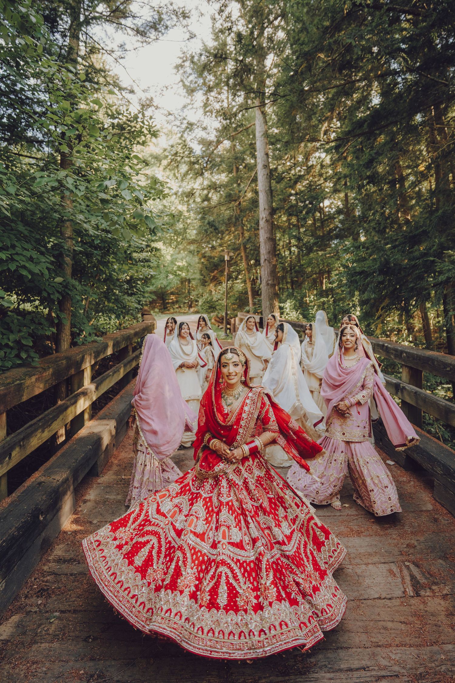Wedding party in red and pink traditional Indian attire posed on wooden bridge in forest setting.