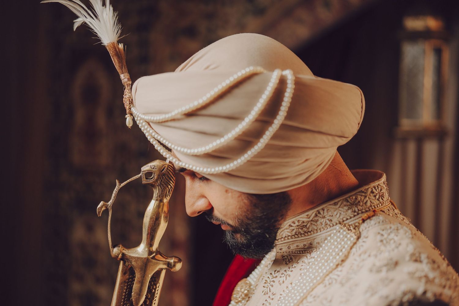 Close-up profile of groom in cream turban and sherwani holding ceremonial sword.