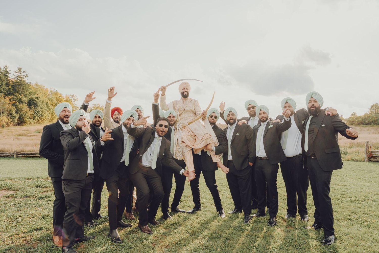 Wedding party members in gray suits and dresses jump joyfully in a line against a scenic outdoor backdrop.