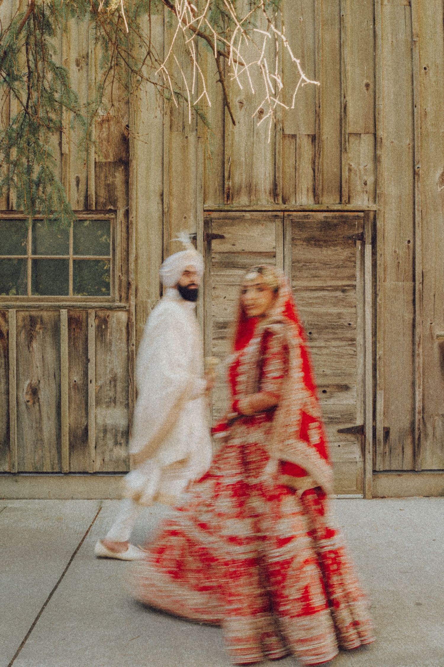 Motion blur shot of couple in traditional red and white attire walking past rustic wooden walls.