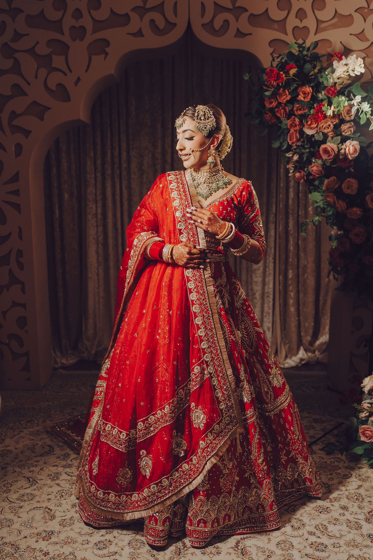 A bride wearing a heavily embellished red lehenga stands gracefully in front of a decorative backdrop.