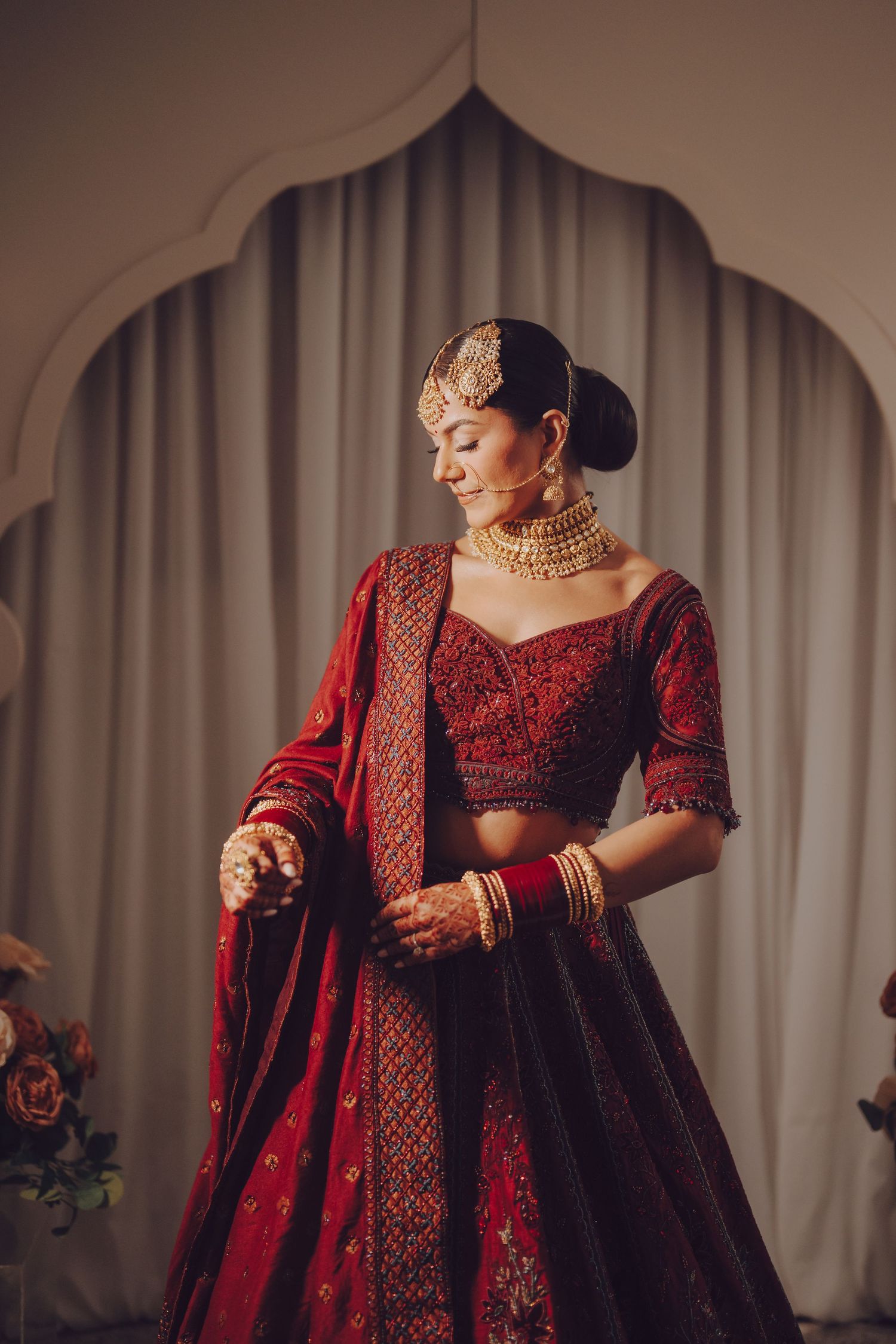 Elegant bride in deep red embroidered lehenga and gold jewelry poses against white arched backdrop.