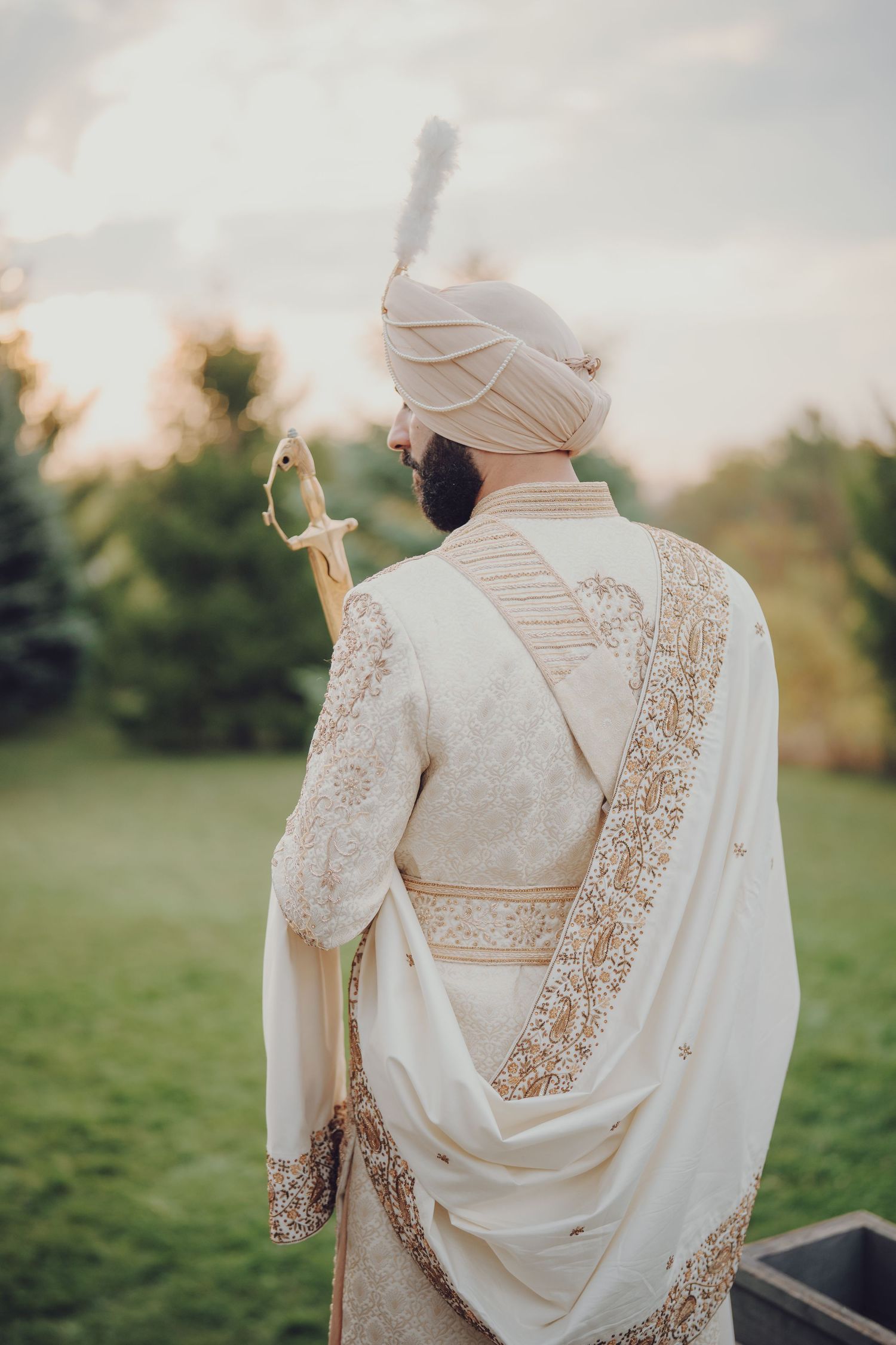 Back view of groom in white and gold embroidered Sikh wedding attire against outdoor setting.