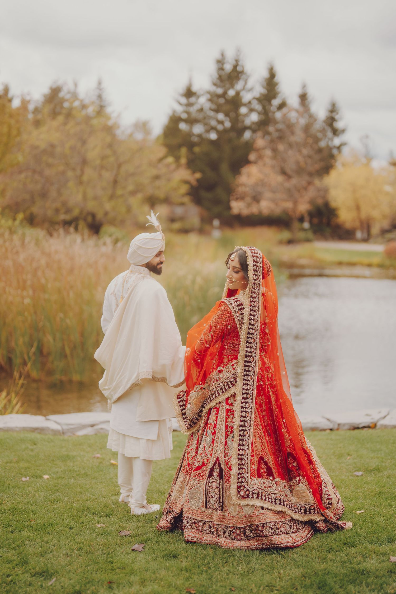Wedding couple in traditional Indian attire poses by lakeside in autumn setting.