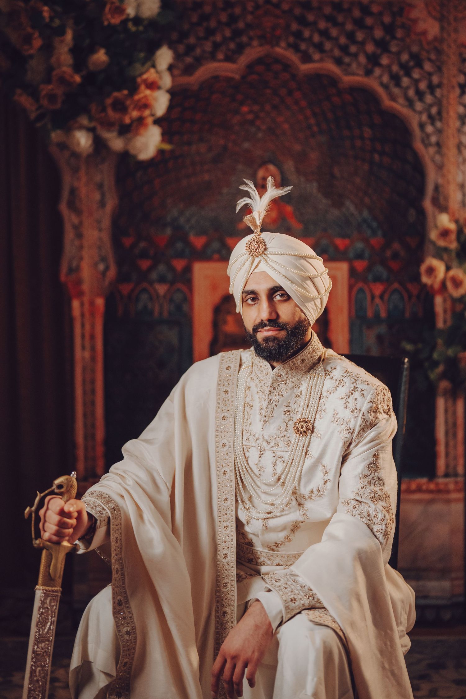 Sikh groom in regal white attire and turban poses with sword against ornate backdrop.