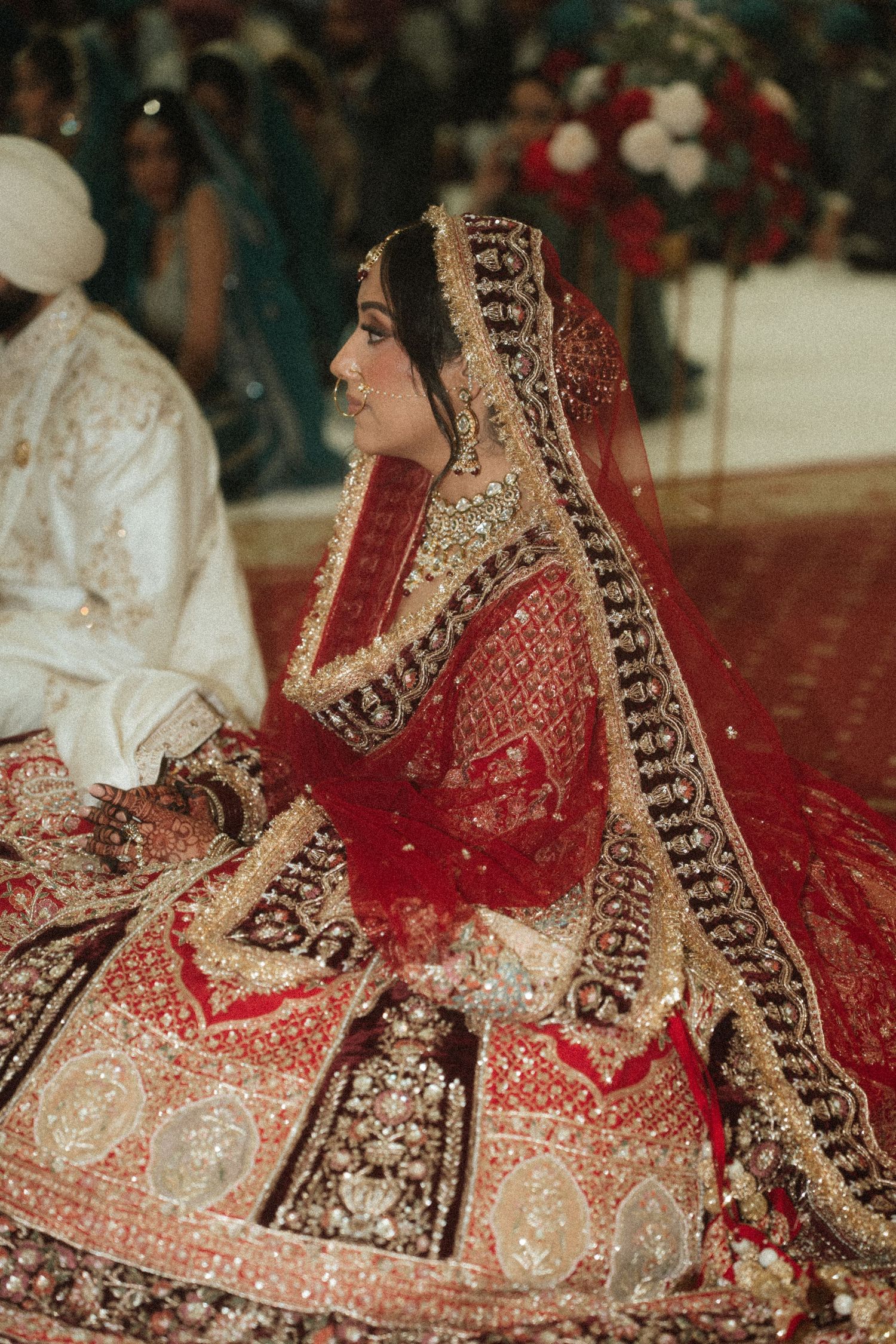 A bride wearing an intricately embroidered red and gold lehenga with detailed border work during a traditional ceremony.